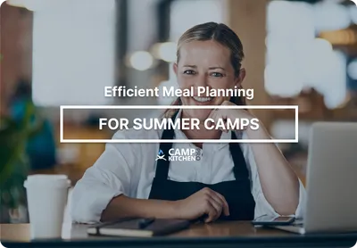 A camp food service manager in an apron planning menus on a laptop at a kitchen workstation.